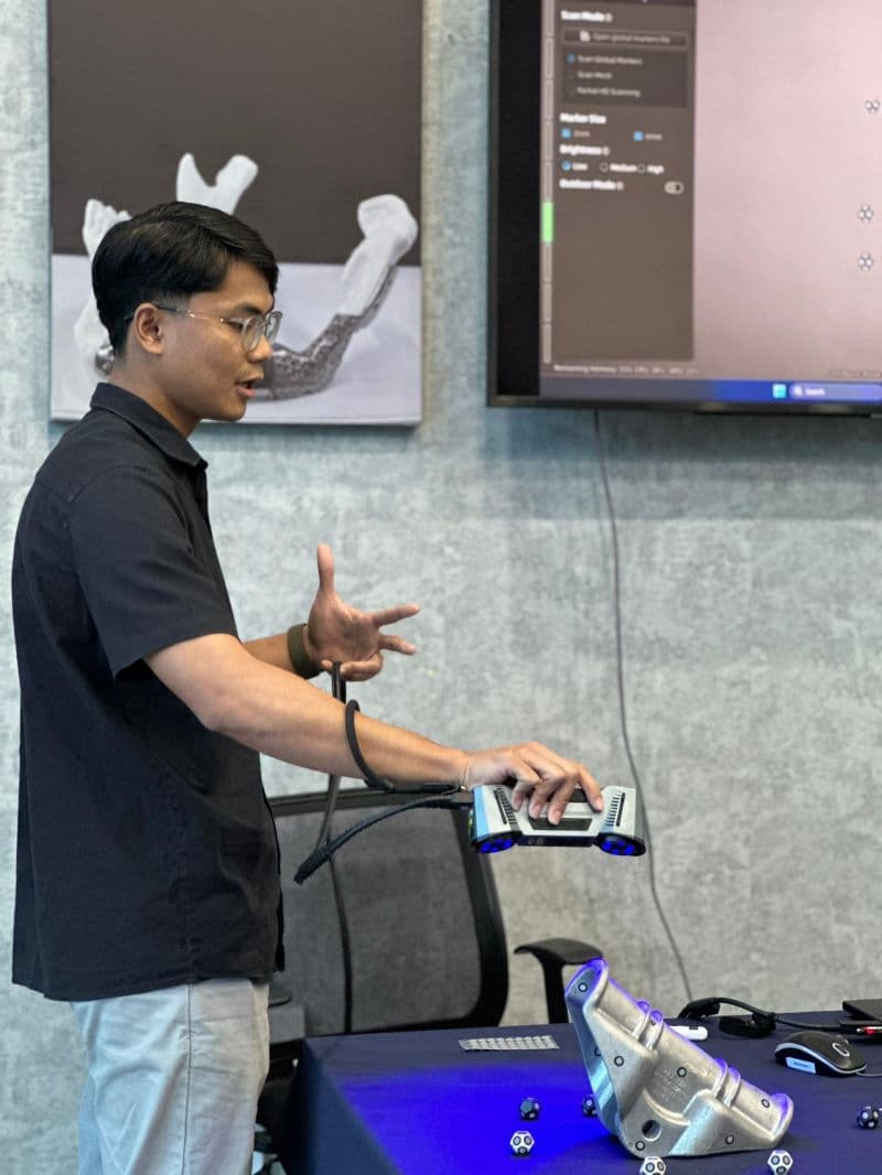 A 3D Gens engineer demonstrates a handheld 3D scanner to the IMMaP UiTM delegation, with a metal-printed part on the table and scan data displayed on the screen behind
